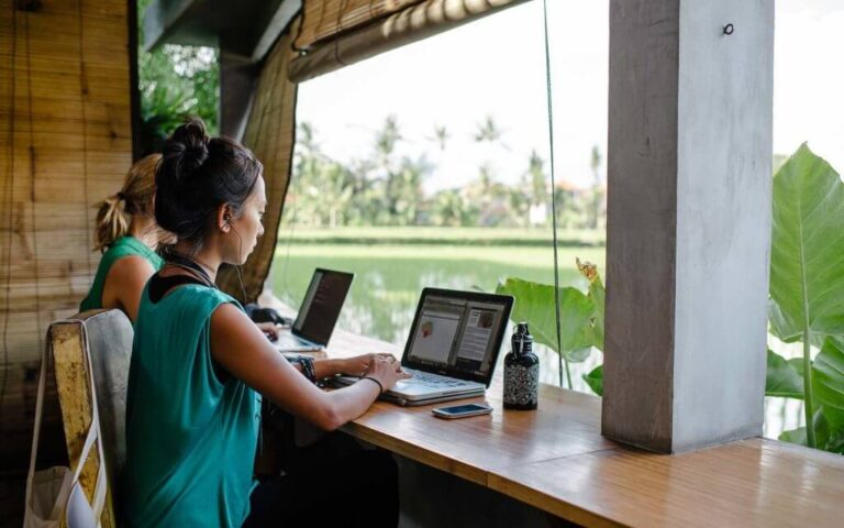 Two digital nomads working remotely on laptops at a coworking space with a tropical rice field view